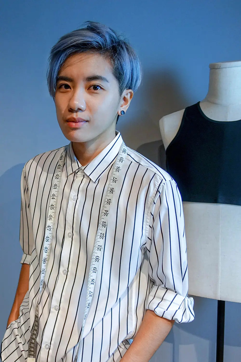 Close-up of a tomboy with striking blue-dyed hair, wearing black stud earrings and a crisp, vertically striped shirt. Behind him, a TOMSCOUT chest binder is showcased on a mannequin, emphasizing inclusive fashion and body-affirming style.