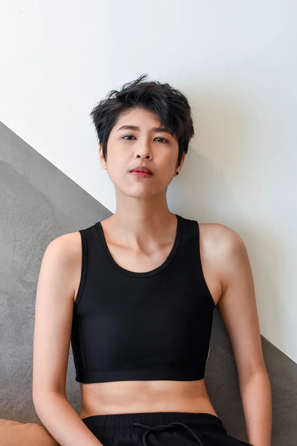 A trans man with textured, short-cropped hair leans confidently against a geometric concrete wall while wearing a sleek, form-fitting black TOMSCOUT chest binder and matching shorts, embodying minimalist style and self-empowerment.