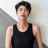 A trans man with textured, short-cropped hair leans confidently against a geometric concrete wall while wearing a sleek, form-fitting black TOMSCOUT chest binder and matching shorts, embodying minimalist style and self-empowerment.