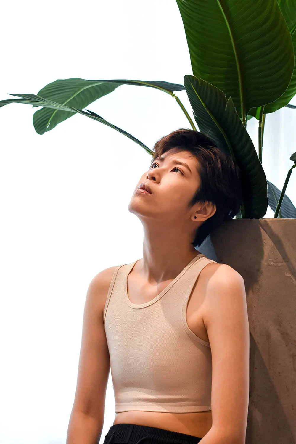 A non-binary individual gazes skyward beneath vibrant tropical leaves, wearing TOMSCOUT’s beige compression binder, showcasing its seamless fit and breathable material for empowered self-expression.