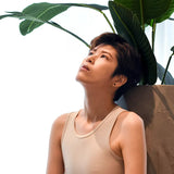 A non-binary individual gazes skyward beneath vibrant tropical leaves, wearing TOMSCOUT’s beige compression binder, showcasing its seamless fit and breathable material for empowered self-expression.