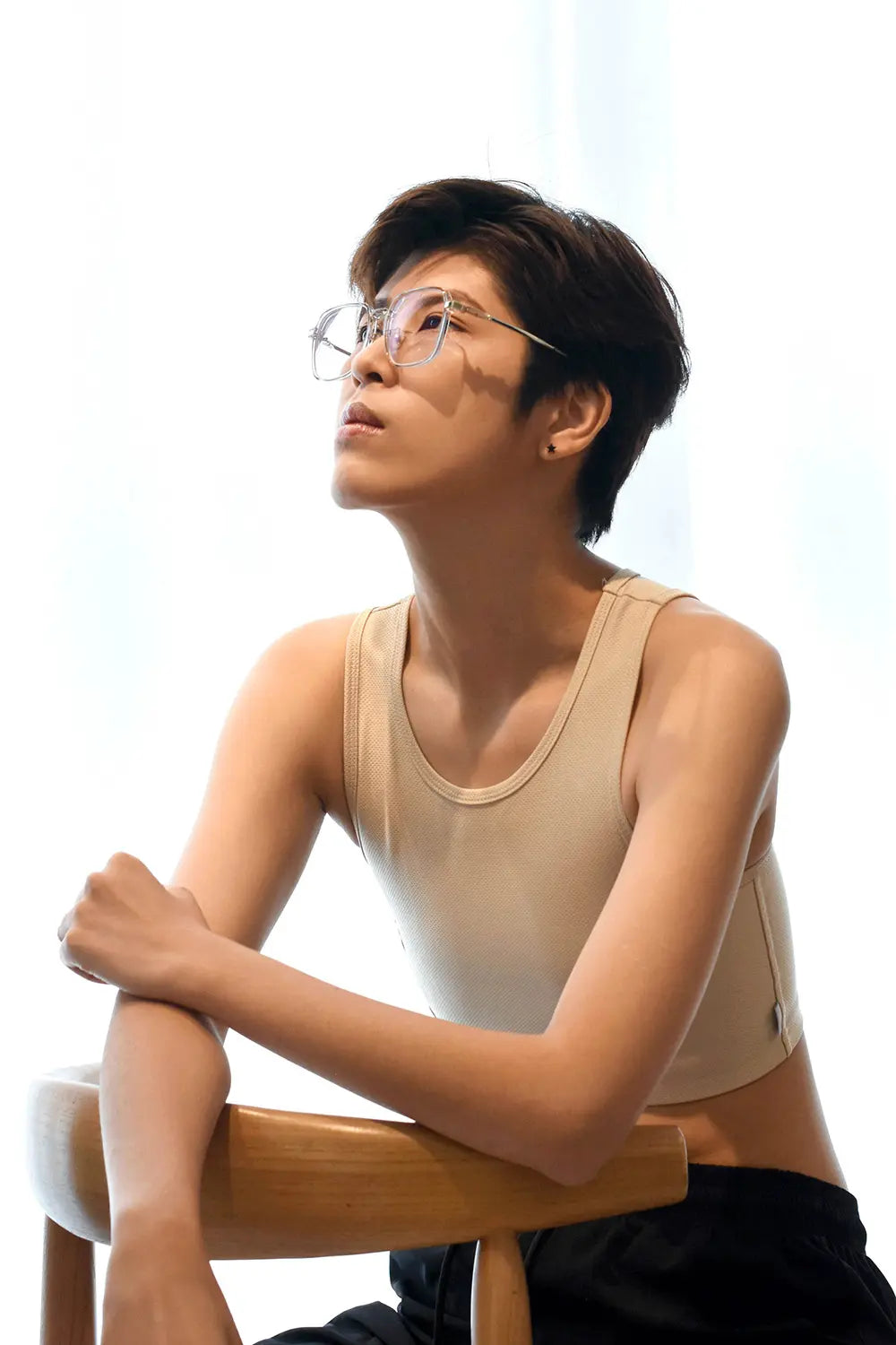 A tomboy with textured short hair poses thoughtfully against a white backdrop, wearing a light beige TOMSCOUT binder that showcases a confident silhouette and minimalist style.
