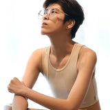A tomboy with textured short hair poses thoughtfully against a white backdrop, wearing a light beige TOMSCOUT binder that showcases a confident silhouette and minimalist style.