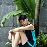 Trans man sits barefoot against lush tropical foliage, arms wrapped around their knees, wearing a TOMSCOUT swim binder and compression shorts—embodying gender euphoria, freedom, and tropical calm.