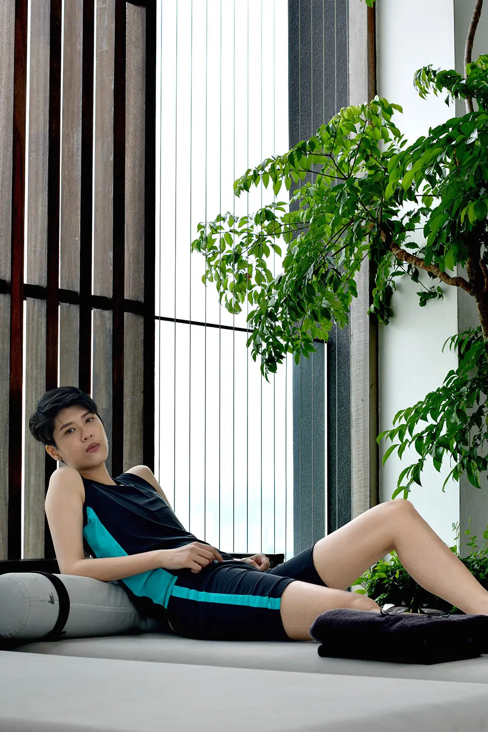 Lesbian model lounges on a sleek daybed in a modern zen-like resort, wearing TOMSCOUT’s gender-affirming swimwear set that combines sporty design with confidence-boosting compression.