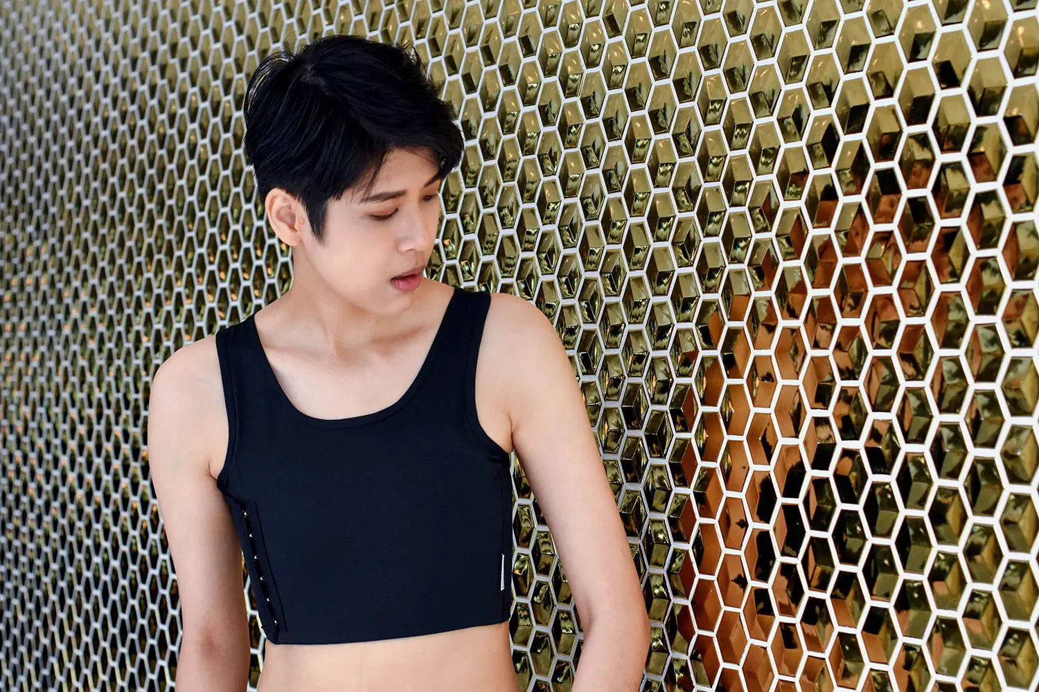 A stylish androgynous model stands before a honeycomb-patterned wall in a matte black TOMSCOUT compression binder, radiating confidence and body autonomy in a structured, masculine silhouette.