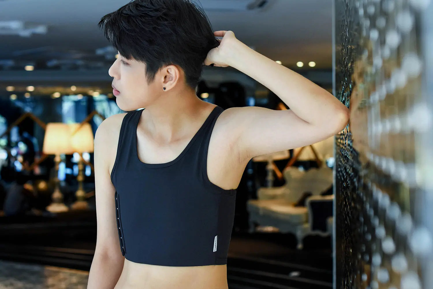 An FTM individual stands against a mirrored wall, gazing outward in a form-fitting TOMSCOUT chest binder—highlighting strong posture and gender-affirming confidence in a chic, athletic cut.