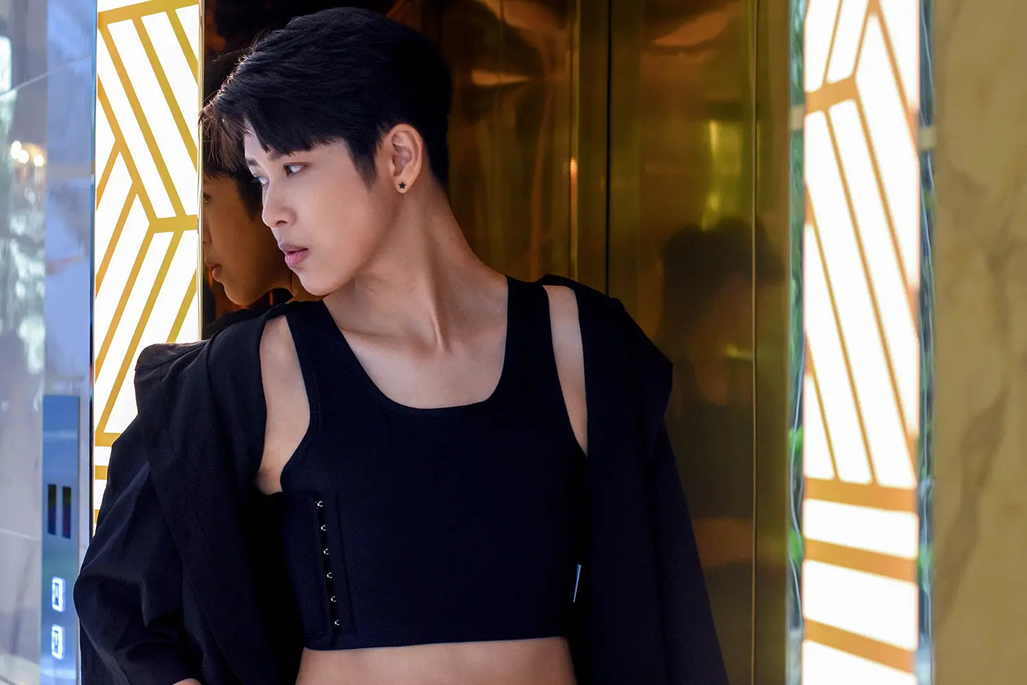 A trans man with short black hair confidently leans against a gold-toned wall in a sleek TOMSCOUT chest binder, featuring side hook detailing for adjustable compression—perfect for daily wear with a bold, minimal look.