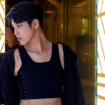 A trans man with short black hair confidently leans against a gold-toned wall in a sleek TOMSCOUT chest binder, featuring side hook detailing for adjustable compression—perfect for daily wear with a bold, minimal look.