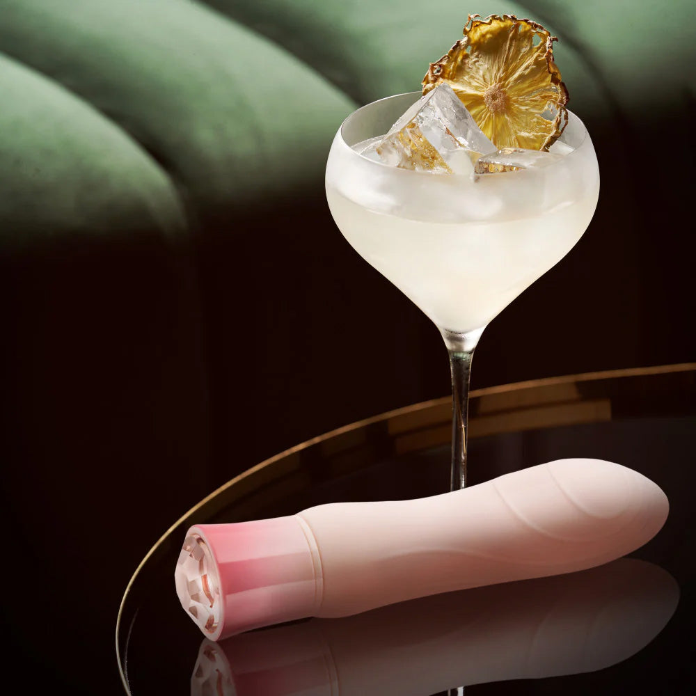 Singapore Blush Oh My Gem rechargeable vibrator placed beside a crystal coupe cocktail with ice and dried pineapple, styled on a luxe velvet surface—fusing elegance and femme-forward pleasure for lesbian and non-binary users.