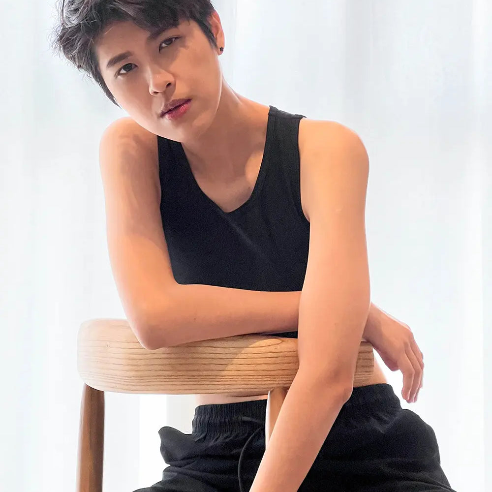 A confident FTM model with tousled short hair reclines against a wooden chair, wearing TOMSCOUT’s sleek black compression binder and matching shorts, exuding cool confidence and androgynous allure.