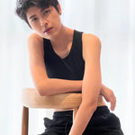 A confident FTM model with tousled short hair reclines against a wooden chair, wearing TOMSCOUT’s sleek black compression binder and matching shorts, exuding cool confidence and androgynous allure.