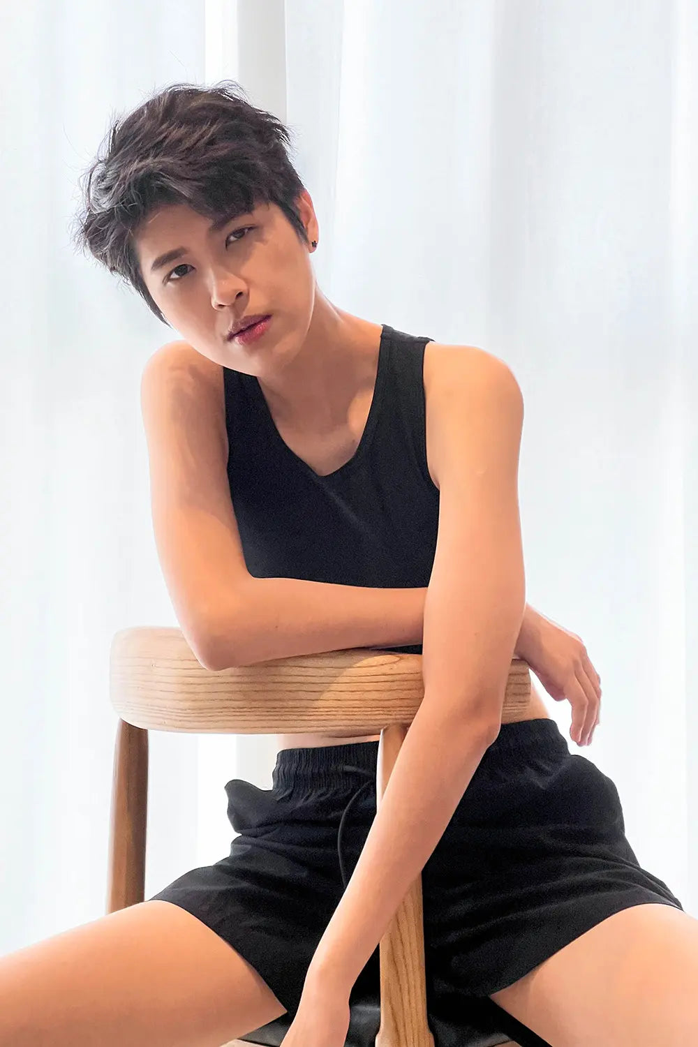 A confident FTM model with tousled short hair reclines against a wooden chair, wearing TOMSCOUT’s sleek black compression binder and matching shorts, exuding cool confidence and androgynous allure.