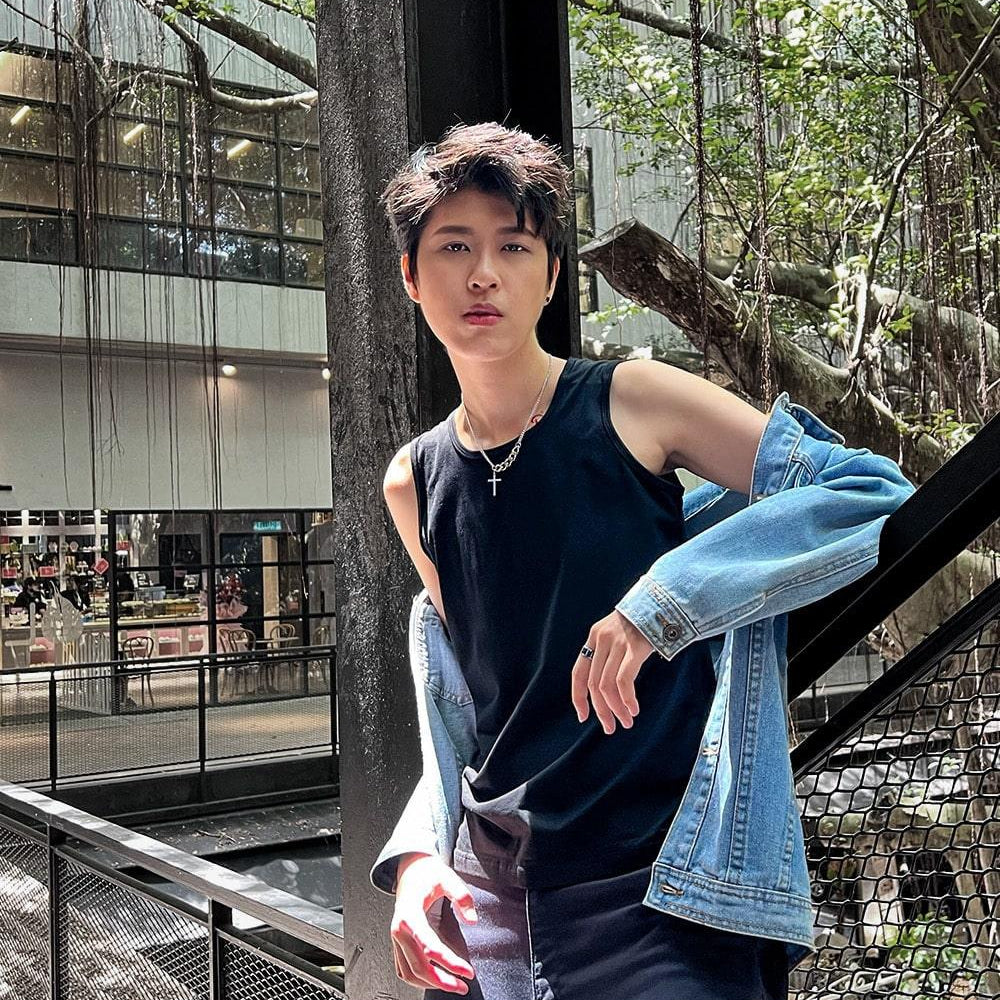 Trans masc-presenting model stands poised against a metal railing, wearing TOMSCOUT’s gender-affirming black chest binder and rugged navy cargo pants, blending street style with soft rebellion in a lush, open-air setting.