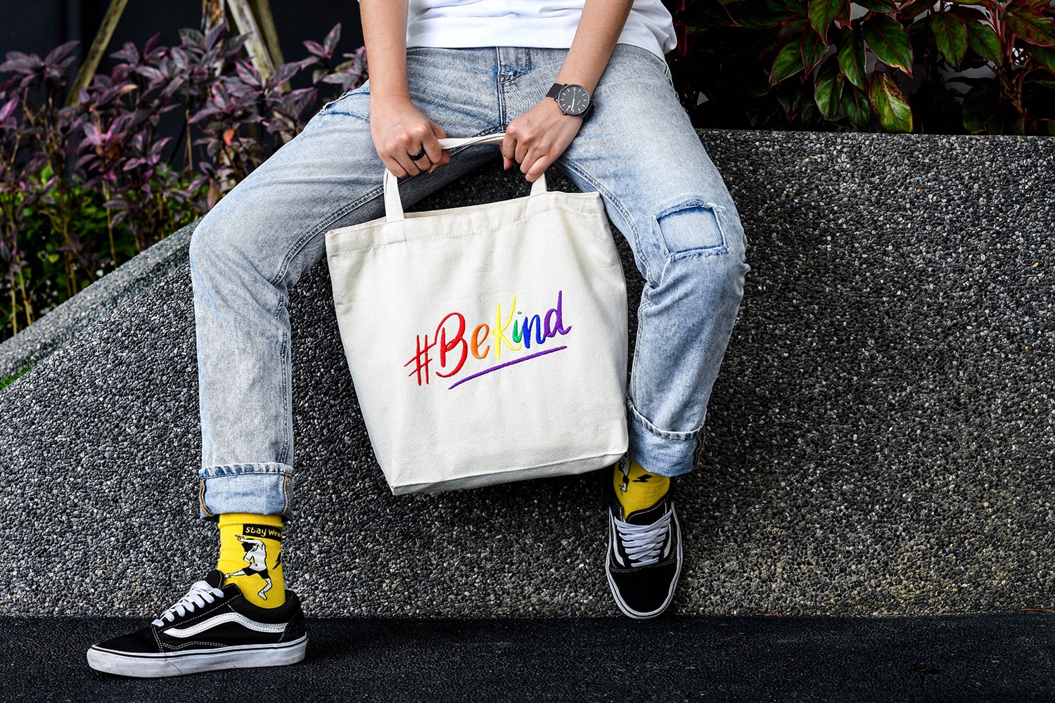 A non-binary androgynous tomboy, transgender man holding a white color embroidered Be Kind canvas tote bag with the LGBTQ+ pride rainbow color design. TOMSCOUT BE KIND - Tote Bag