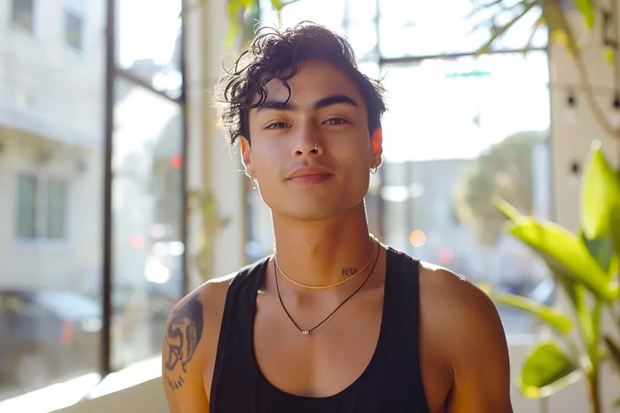 Confident FTM individual with tousled curls and minimalist jewelry poses in natural sunlight wearing a sleek black TOMSCOUT chest binder, showcasing gender-affirming style and empowered self-expression. The soft, breathable fabric hugs his frame comfortably, perfect for everyday wear or layering. Sunlight streams through urban studio windows, highlighting tattoos and body confidence.