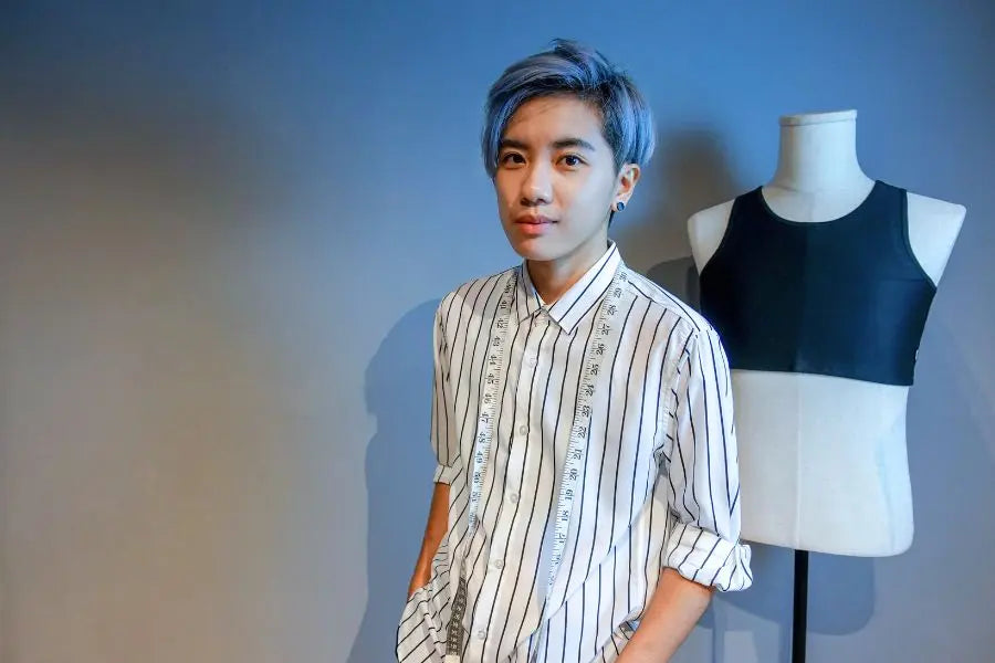 Androgynous fashion designer with lavender hair and a striped button-up shirt stands confidently beside a mannequin wearing a black TOMSCOUT chest binder. Draped with a measuring tape, they embody queer creativity and precision tailoring, showcasing TOMSCOUT’s commitment to gender-affirming design that fuses functionality, identity, and inclusive style.