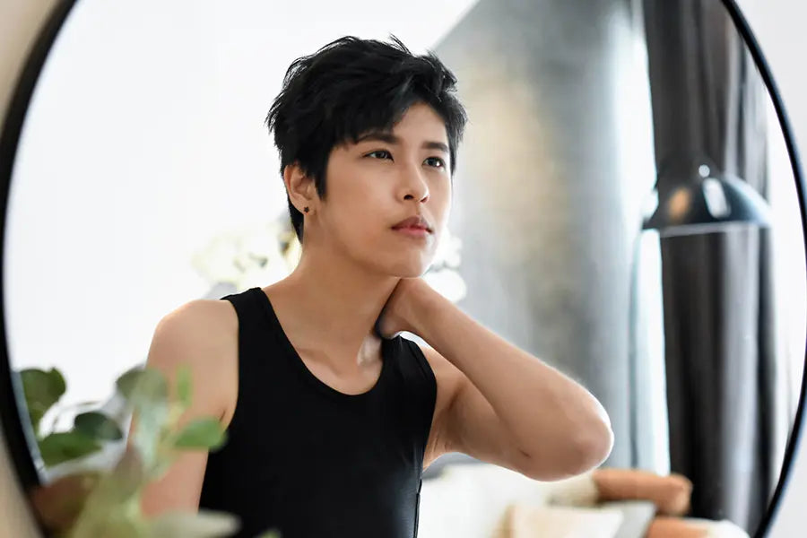 Androgynous individual with tousled short hair gazes into a round mirror while wearing a form-fitting black TOMSCOUT chest binder, reflecting on self-identity and body alignment. The soft lighting and minimalist bedroom decor evoke a calm, introspective mood, capturing the empowering ritual of daily gender affirmation.