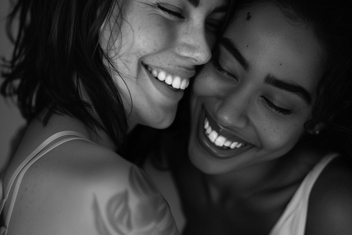 Intimate black and white portrait of two femmes laughing with their foreheads touching—capturing soft queer joy, lesbian love, and the tenderness of LGBTQ+ connection.