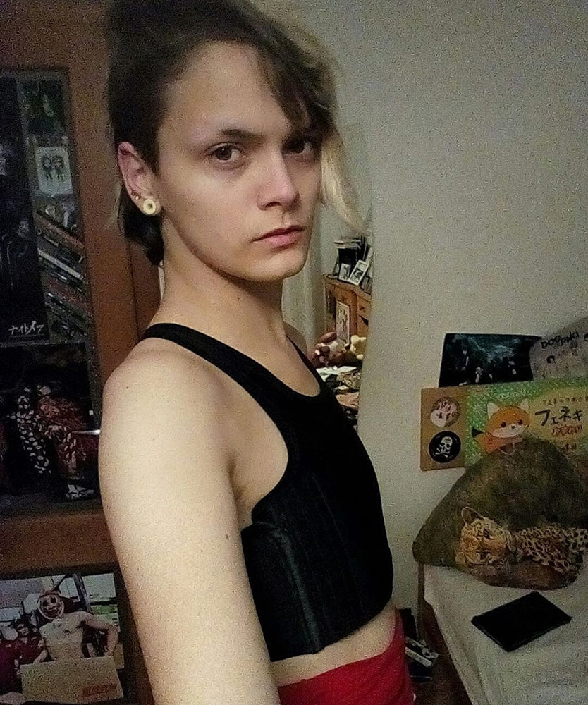 A non-binary person with a half-shaved hairstyle poses in a black TOMSCOUT binder, looking directly at the camera with a calm, self-assured expression—sharing a testimonial about comfort, support, and identity validation.