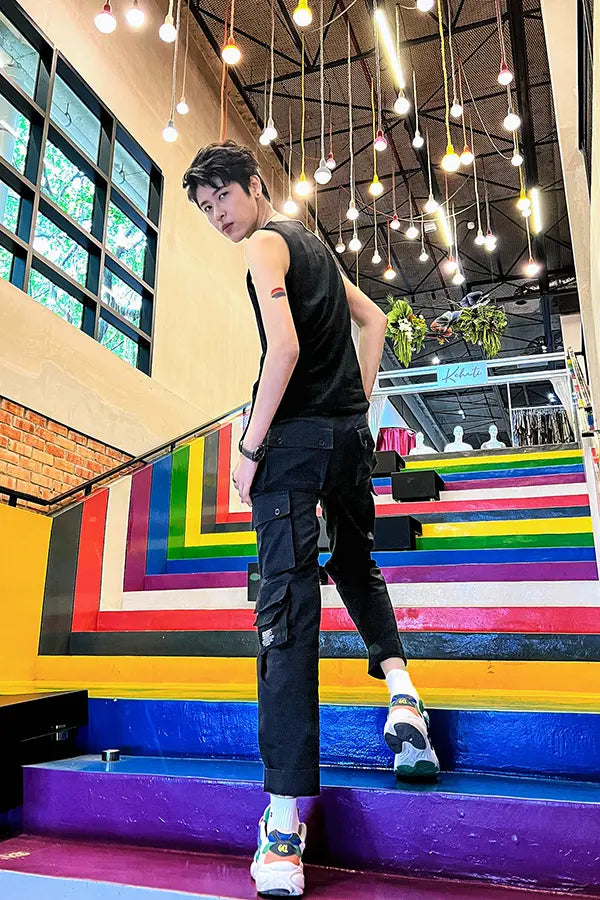 FTM model with a rainbow pride tattoo on his arm stands boldly on vibrant rainbow-painted stairs, wearing a black TOMSCOUT chest binder, black cargo pants, and chunky sneakers, illuminated by a canopy of hanging lights, representing fearless gender expression and queer celebration.