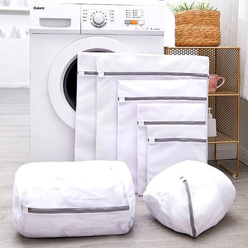 Set of mesh laundry bags in various sizes arranged beside a front-loading washing machine. These breathable zippered wash bags are ideal for preserving TOMSCOUT binders, boxer briefs, and delicate gender-affirming wear.