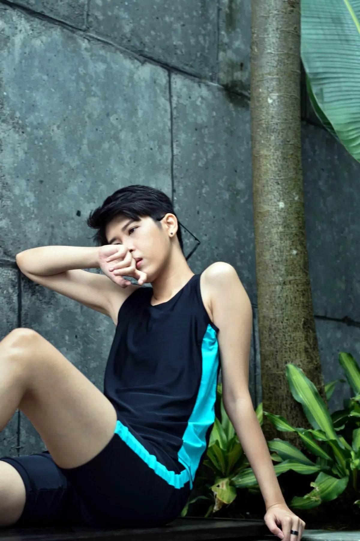 Androgynous non-binary person in a sapphire-accented swim binder, sitting thoughtfully in a quiet outdoor setting — gender-affirming TOMSCOUT swimwear for transmasc and lesbian identities.