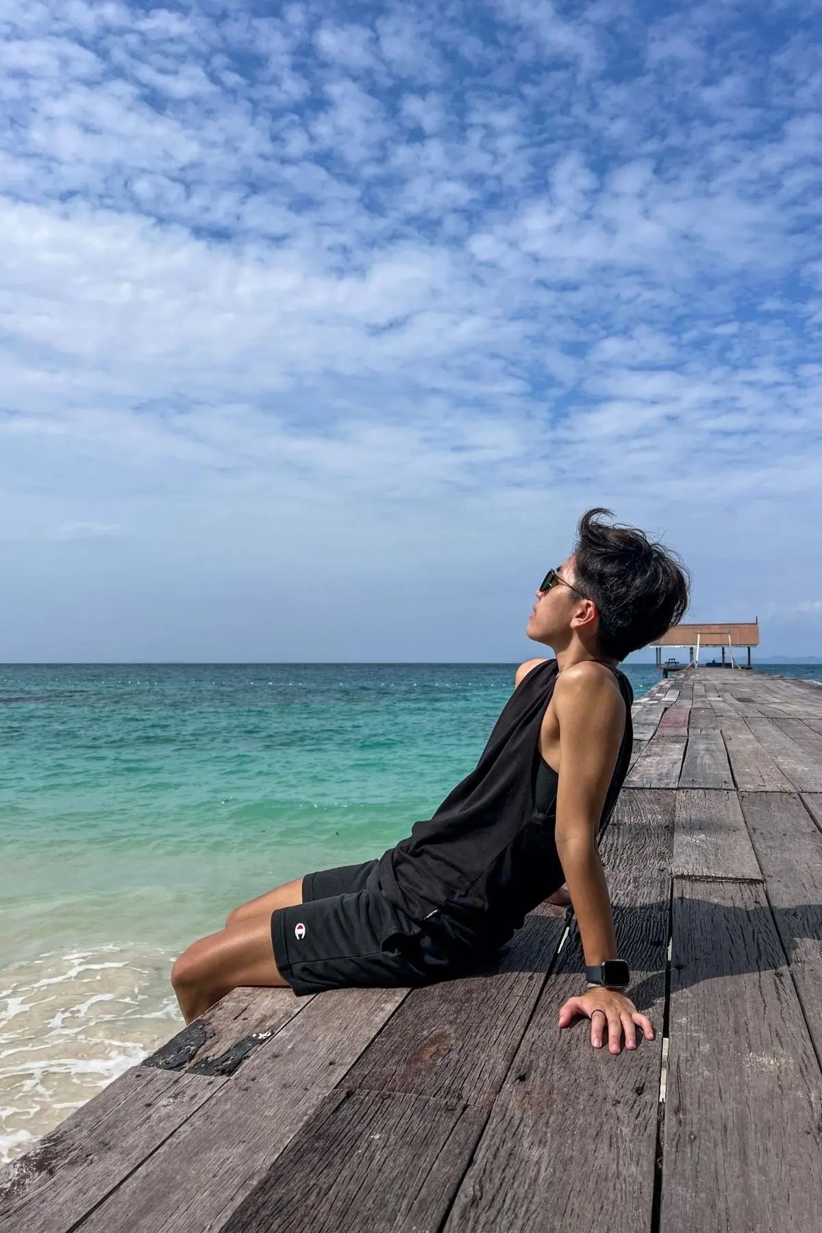 Transmasc person in a TOMSCOUT black swim binder set sitting confidently on a tropical dock, gazing at the sea — swimwear for FTM transgender and tomboy swimmers.