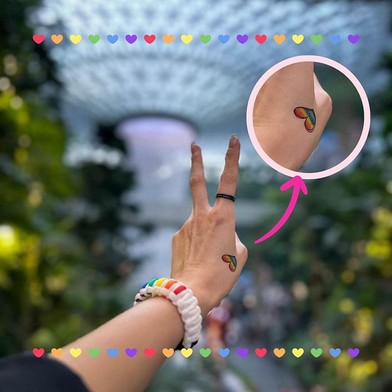 Hand flashing a peace sign with a rainbow heart tattoo on the wrist, surrounded by vibrant mini hearts—queer joy and Singapore pride captured in a TOMSCOUT tattoo sticker moment.