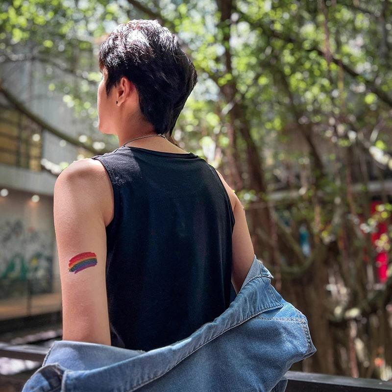 FTM-presenting person showing off a rainbow pride tattoo on their upper arm while wearing a sleeveless black top—TOMSCOUT’s unapologetic celebration of trans pride and outdoor confidence.