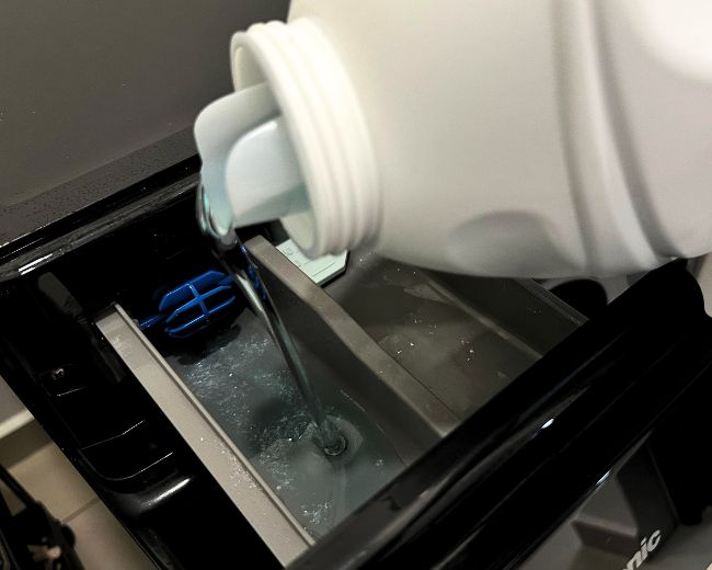 Pouring mild detergent into a washing machine—start of the TOMSCOUT chest binder washing process using gentle, binder-safe detergent.