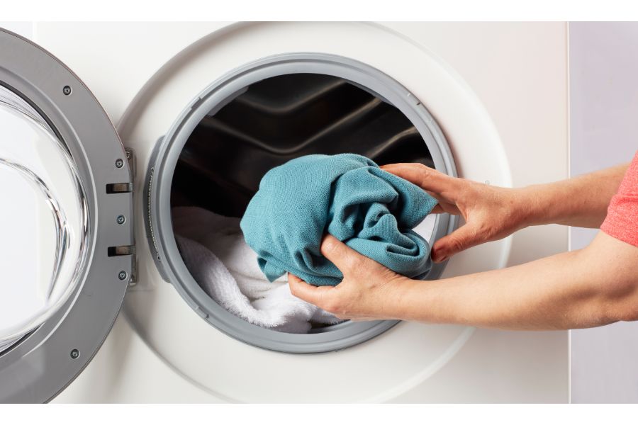 Transgender individual, embodying pride and the essence of the LGBT community, carefully removing their laundry, including a chest binder symbolizing FTM identity, from the washer after a wash cycle, highlighting a moment of domestic life and self-care.