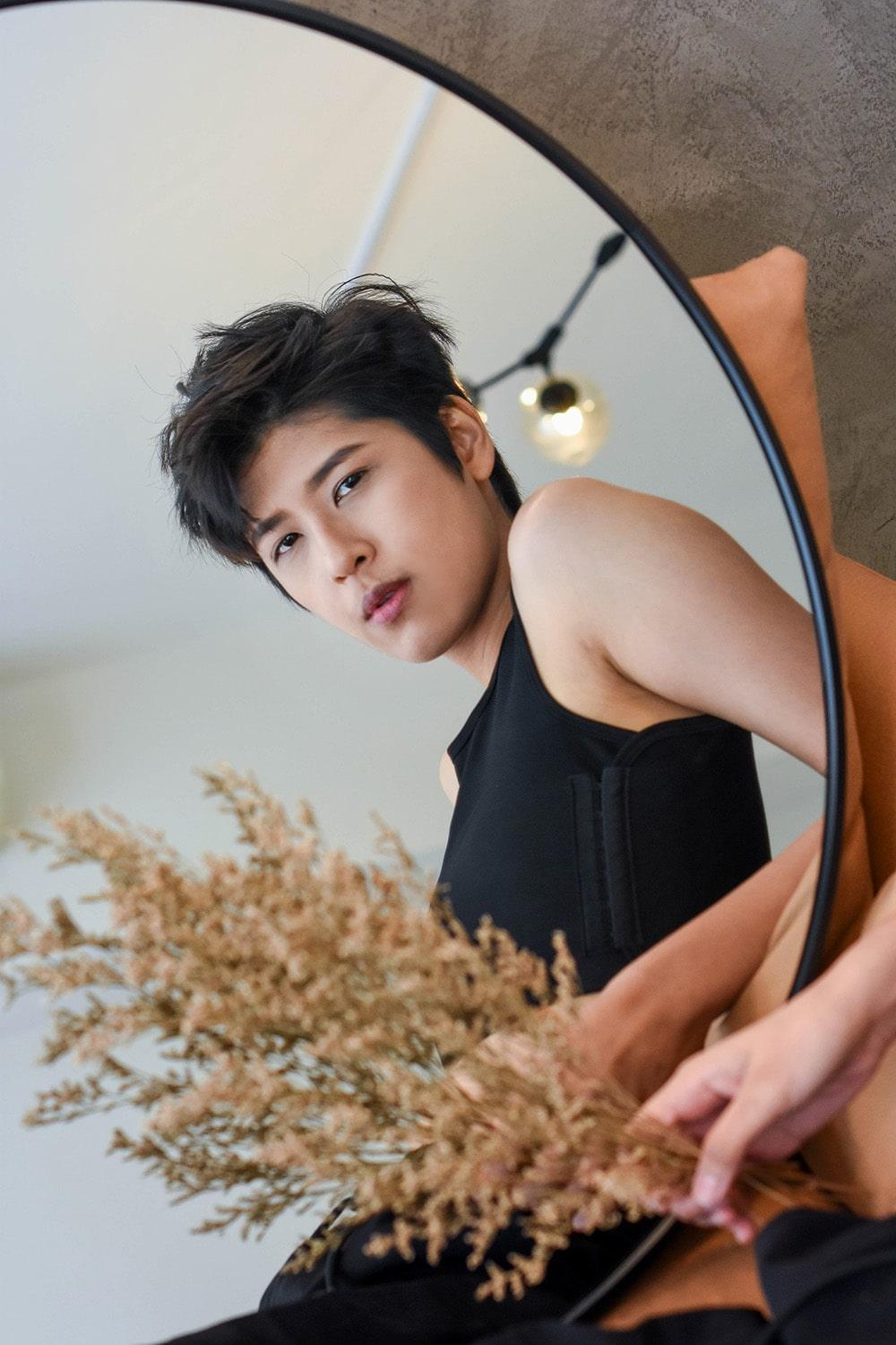 A non-binary androgynous tomboy, transgender man sitting while holding a flower as a prop, dressed in a black non-bandage TOMSCOUT Chest Binder from the BECOMING collection, exuding confidence as they look towards the camera.