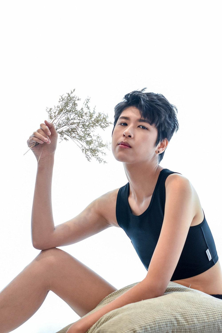 A non-binary androgynous tomboy, transgender man sitting and holding a flower as prop and wearing a black color of a non-bandage TOMSCOUT Chest Binder to fight body dysphoria and looking confidence towards the camera. TOMSCOUT BECOMING non-bandage binder.