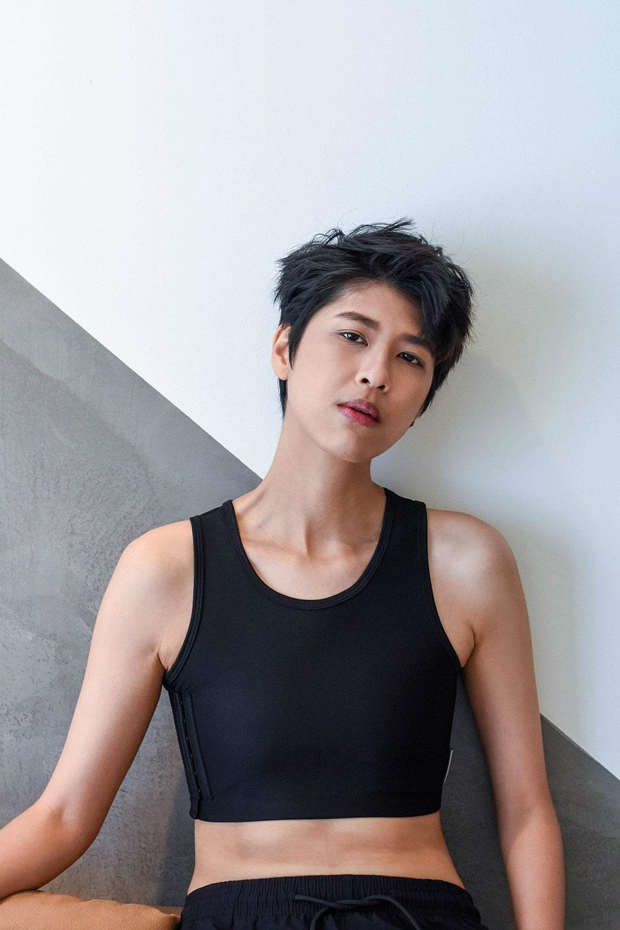 A non-binary androgynous tomboy, transgender man sitting and wearing a black color of a non-bandage TOMSCOUT Chest Binder to fight body dysphoria and looking confidence towards the camera. TOMSCOUT BECOMING non-bandage binder.