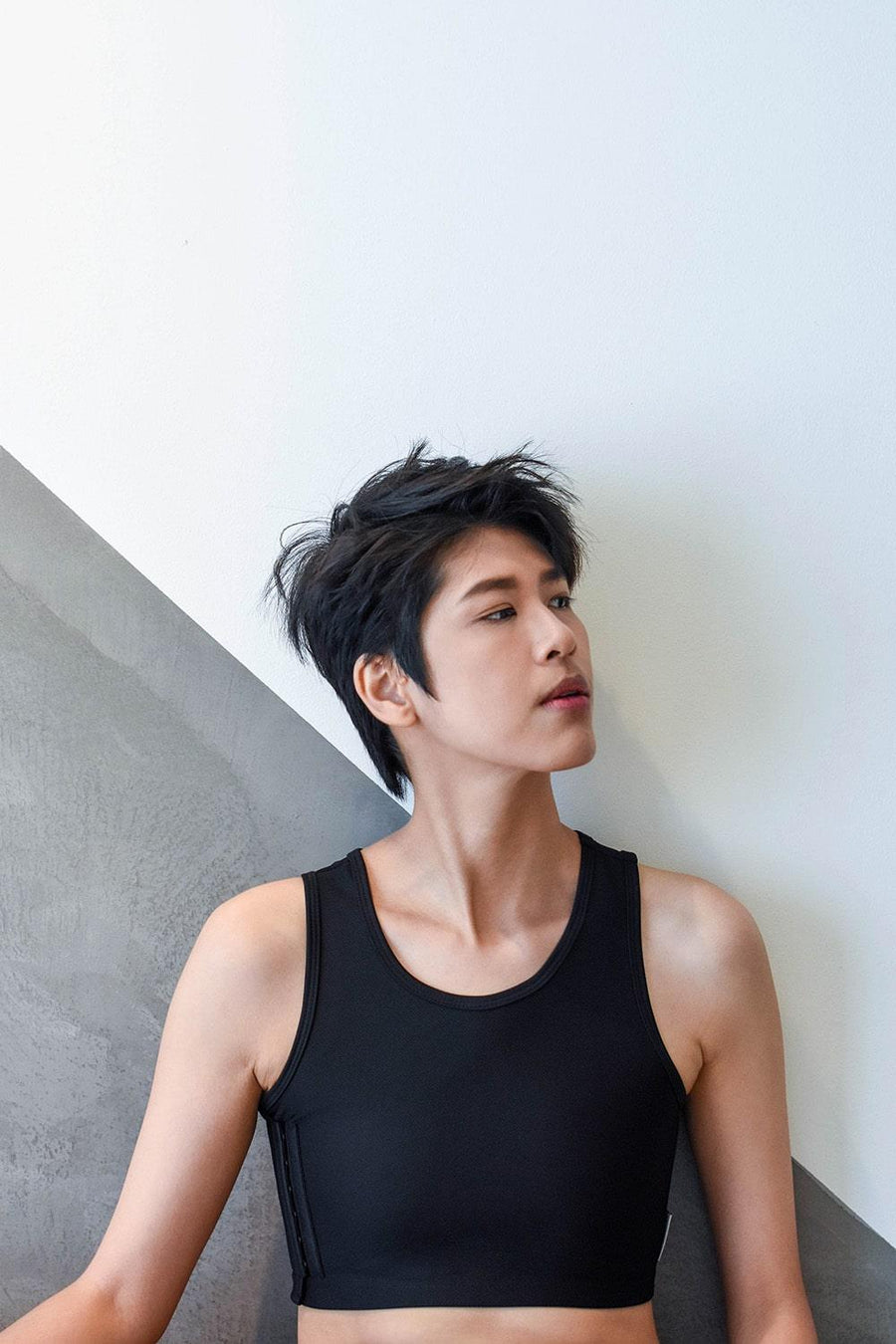 A non-binary androgynous tomboy, transgender man sitting and wearing a black color of a non-bandage TOMSCOUT Chest Binder to fight body dysphoria and looking towards left side. TOMSCOUT BECOMING non-bandage binder.