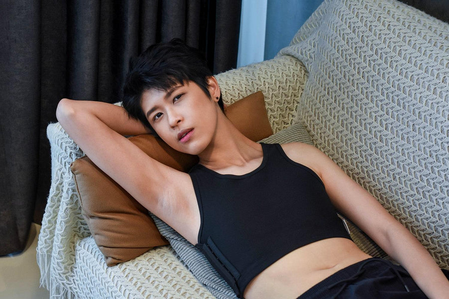 A non-binary androgynous tomboy, transgender man lying on a leather sofa and wearing a black color of a non-bandage TOMSCOUT Chest Binder to fight body dysphoria and looking confidence towards the camera. TOMSCOUT BECOMING non-bandage binder.