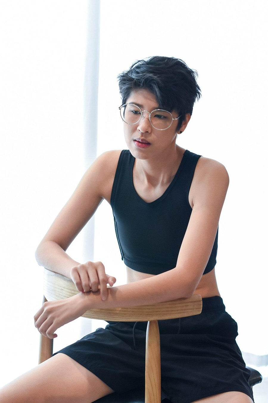 A non-binary androgynous tomboy, transgender man sitting and wearing a black color of a non-bandage TOMSCOUT Chest Binder to fight body dysphoria and looking confidence towards the camera. TOMSCOUT BECOMING non-bandage binder.