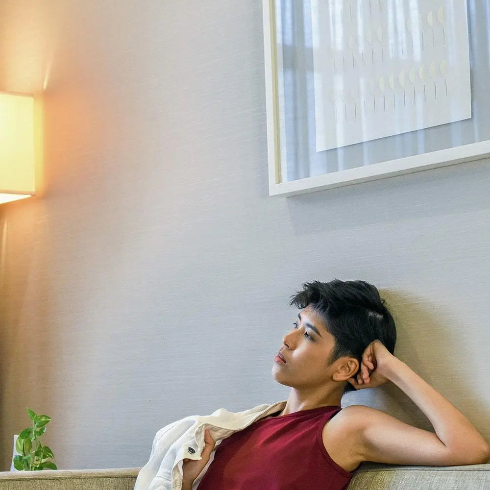 Non-binary individual lounging thoughtfully on a sofa in a maroon TOMSCOUT chest binder tank, radiating self-love and quiet empowerment in a cozy, softly lit living space designed for authenticity and ease.