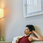 Non-binary individual lounging thoughtfully on a sofa in a maroon TOMSCOUT chest binder tank, radiating self-love and quiet empowerment in a cozy, softly lit living space designed for authenticity and ease.