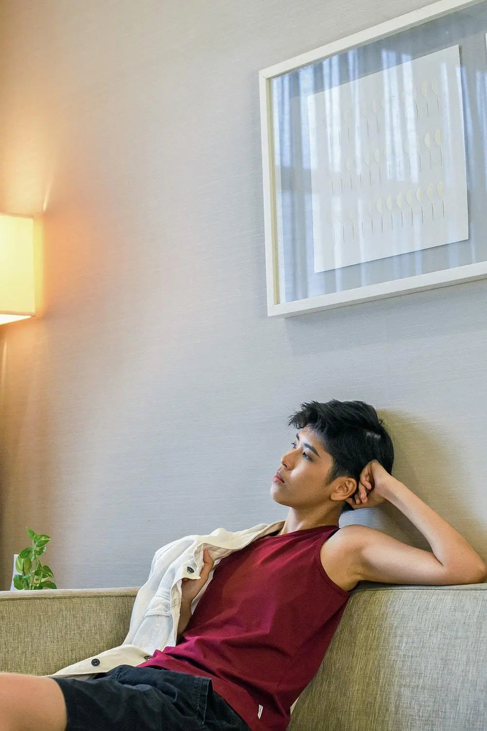 Non-binary individual lounging thoughtfully on a sofa in a maroon TOMSCOUT chest binder tank, radiating self-love and quiet empowerment in a cozy, softly lit living space designed for authenticity and ease.