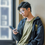 Stylish trans man standing by a sunlit window, focused on his phone while wearing a TOMSCOUT compression tee under an olive green t-shirt and windbreaker—channeling laid-back urban cool with gender euphoria at its core.