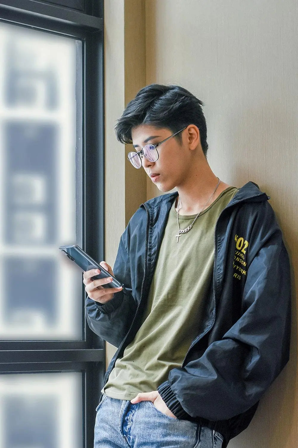 Stylish trans man standing by a sunlit window, focused on his phone while wearing a TOMSCOUT compression tee under an olive green t-shirt and windbreaker—channeling laid-back urban cool with gender euphoria at its core.