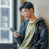 Stylish trans man standing by a sunlit window, focused on his phone while wearing a TOMSCOUT compression tee under an olive green t-shirt and windbreaker—channeling laid-back urban cool with gender euphoria at its core.