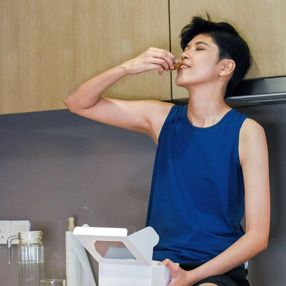 Androgynous person with a confident smile enjoying a sweet treat in a minimalist kitchen, wearing a TOMSCOUT chest binder under a cobalt blue muscle tank—effortlessly blending comfort, gender affirmation, and everyday joy.