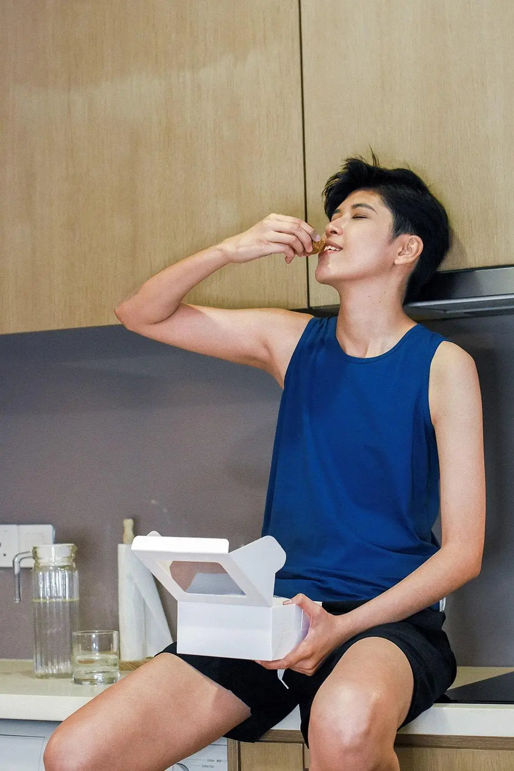 Androgynous person with a confident smile enjoying a sweet treat in a minimalist kitchen, wearing a TOMSCOUT chest binder under a cobalt blue muscle tank—effortlessly blending comfort, gender affirmation, and everyday joy.