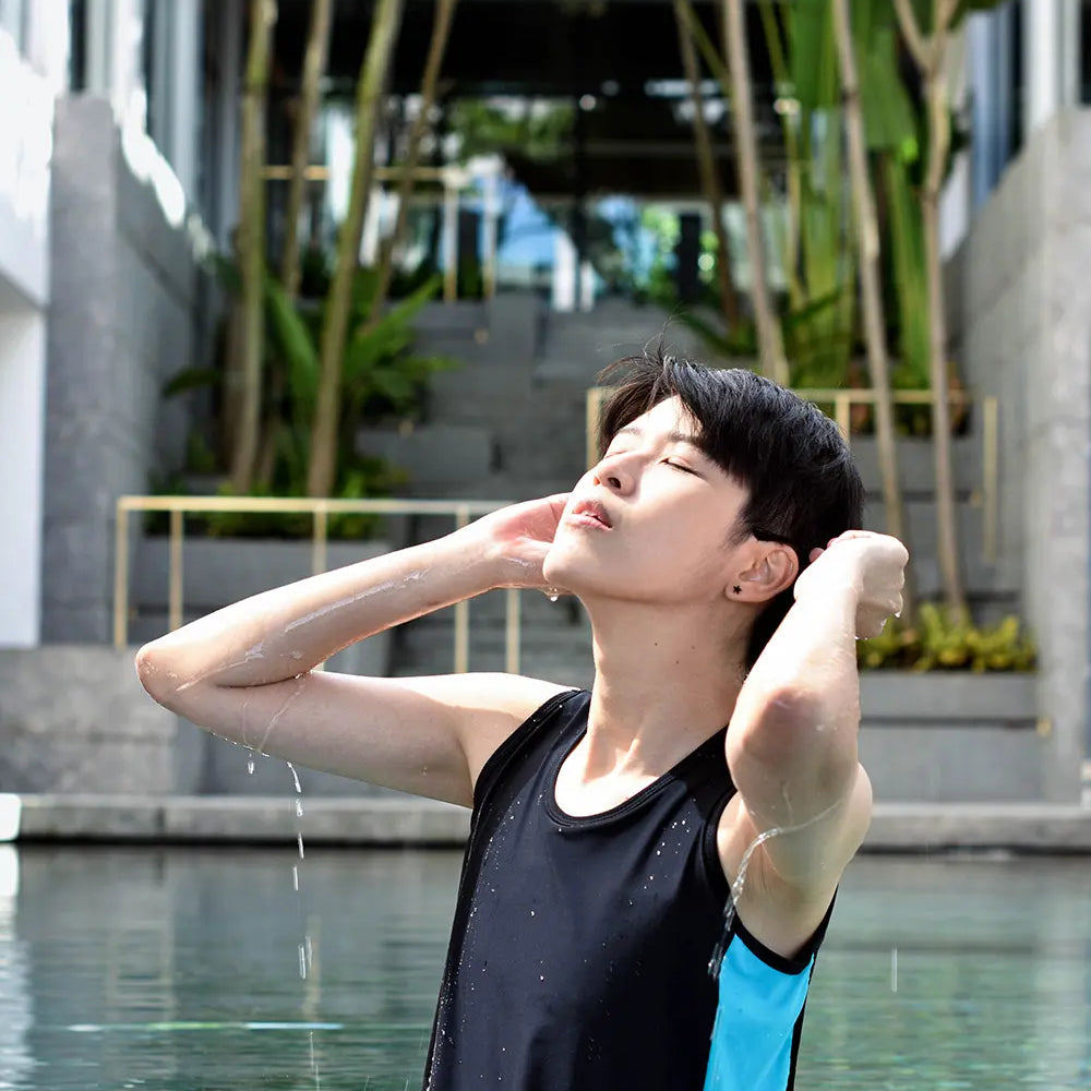 FTM individual emerges confidently from a pool wearing a TOMSCOUT swim binder in black with teal side panels, eyes closed in a moment of self-assured peace, water cascading from their arms under tropical resort light.