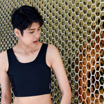 A stylish androgynous model stands before a honeycomb-patterned wall in a matte black TOMSCOUT compression binder, radiating confidence and body autonomy in a structured, masculine silhouette.