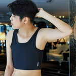 An FTM individual stands against a mirrored wall, gazing outward in a form-fitting TOMSCOUT chest binder—highlighting strong posture and gender-affirming confidence in a chic, athletic cut.
