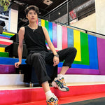 Lesbian-presenting fashionista rocks TOMSCOUT’s compression tank and black cuffed trousers, lounging effortlessly on colorful stairs framed by Progress Pride hues—a radiant tribute to queer joy and self-expression.